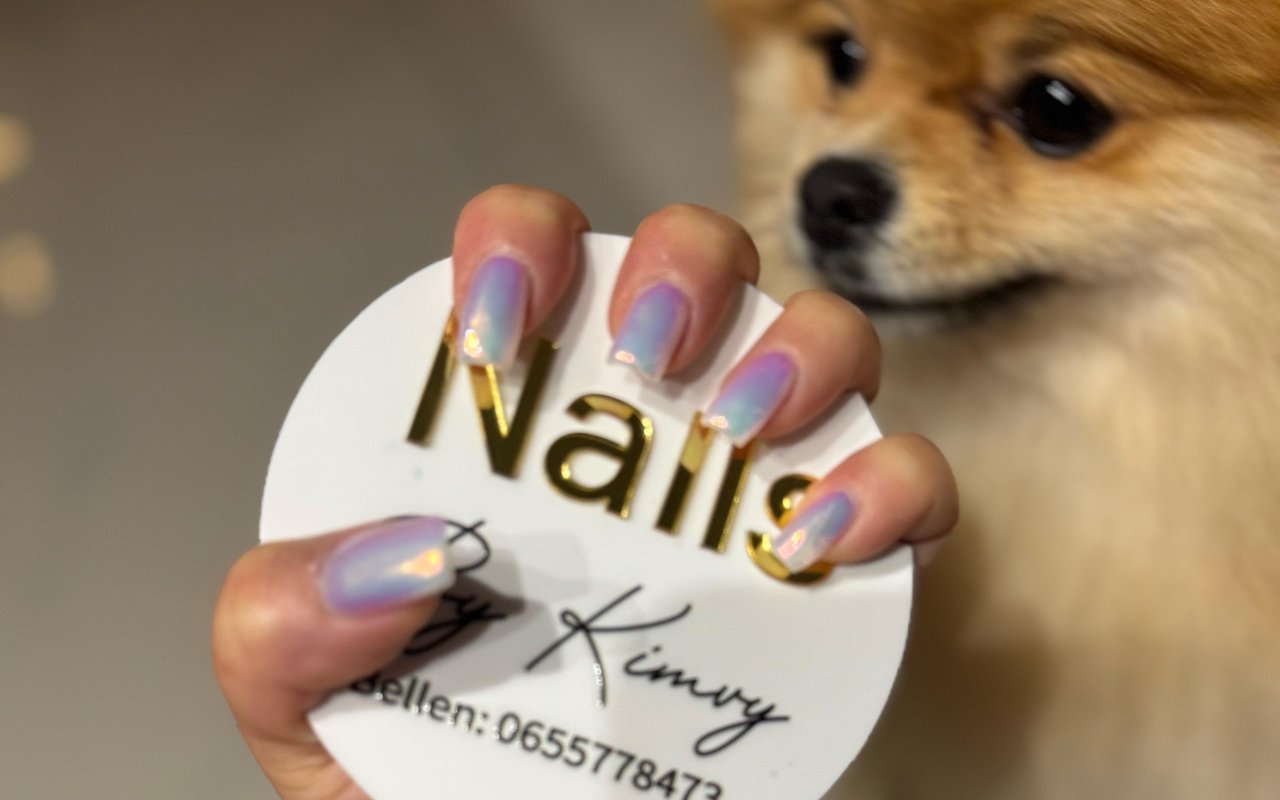 Hand met chroom nagels die het ronde Nails By Kimvy bordje vasthoudt, haar hondje kijkt mee
